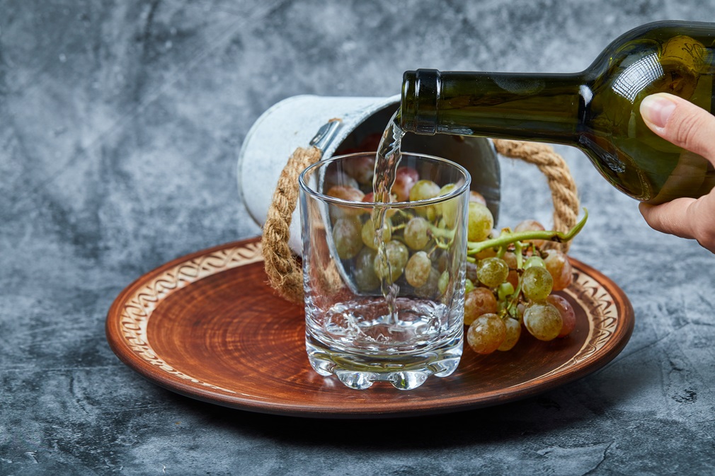 Small bucket of grapes inside ceramic plate and hand pouring wine into the glass on a marble background