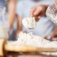Baker tipping wooden spoon full of flour on table surface
