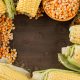 Corn grains with cobs in wooden spoon and plate on wooden background, flat lay.